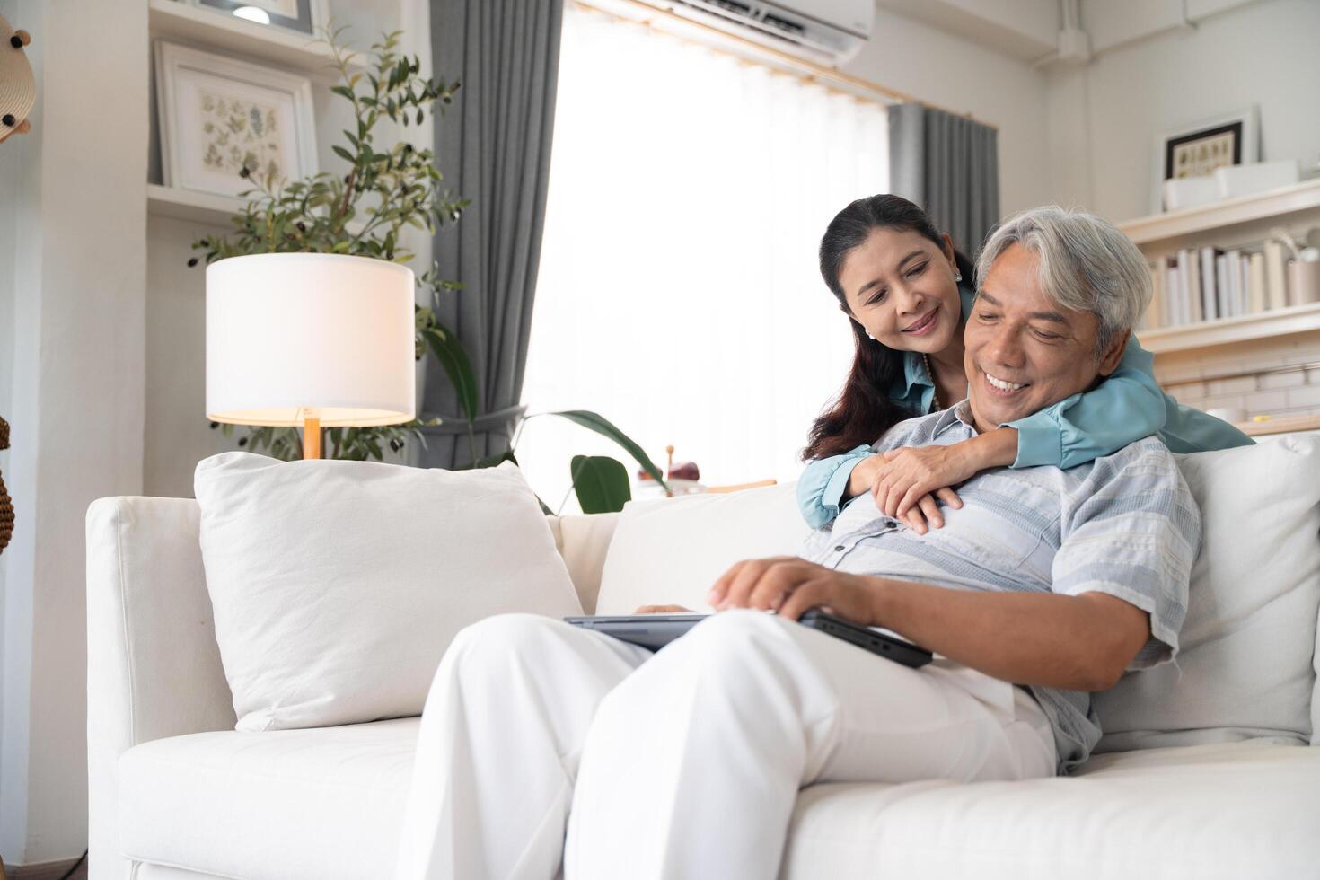 Husband sitting on couch working on laptop while his wife support by embrancing. Elderly couple call with family in the living room at home photo