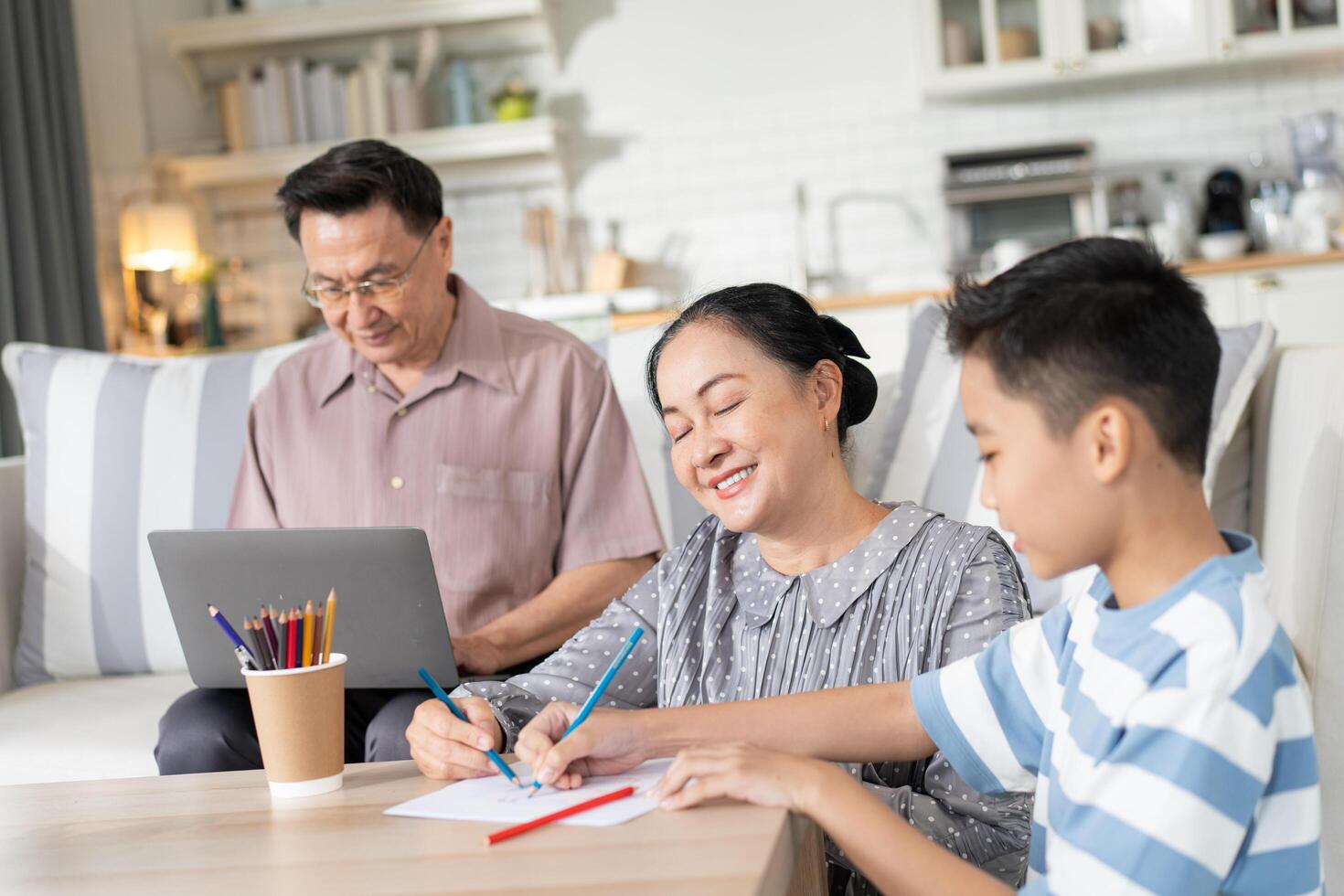 Asian grandmother helping grandson doing homework while grandfather working with laptop at home. Family spending time on weekend together photo