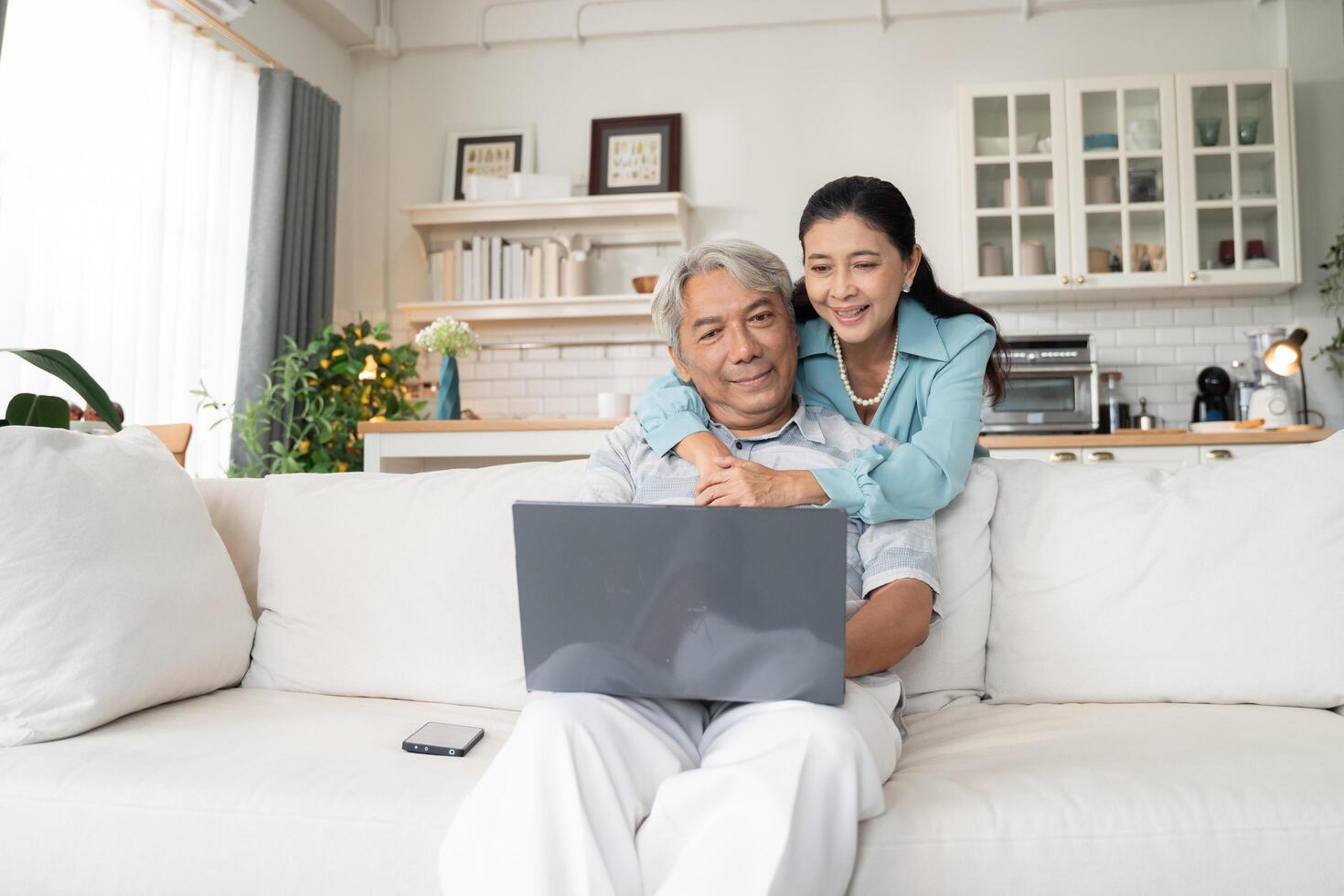 Senior husband sitting on couch working on laptop while his wife support by embrancing. Elderly couple call with family in the living room at home photo