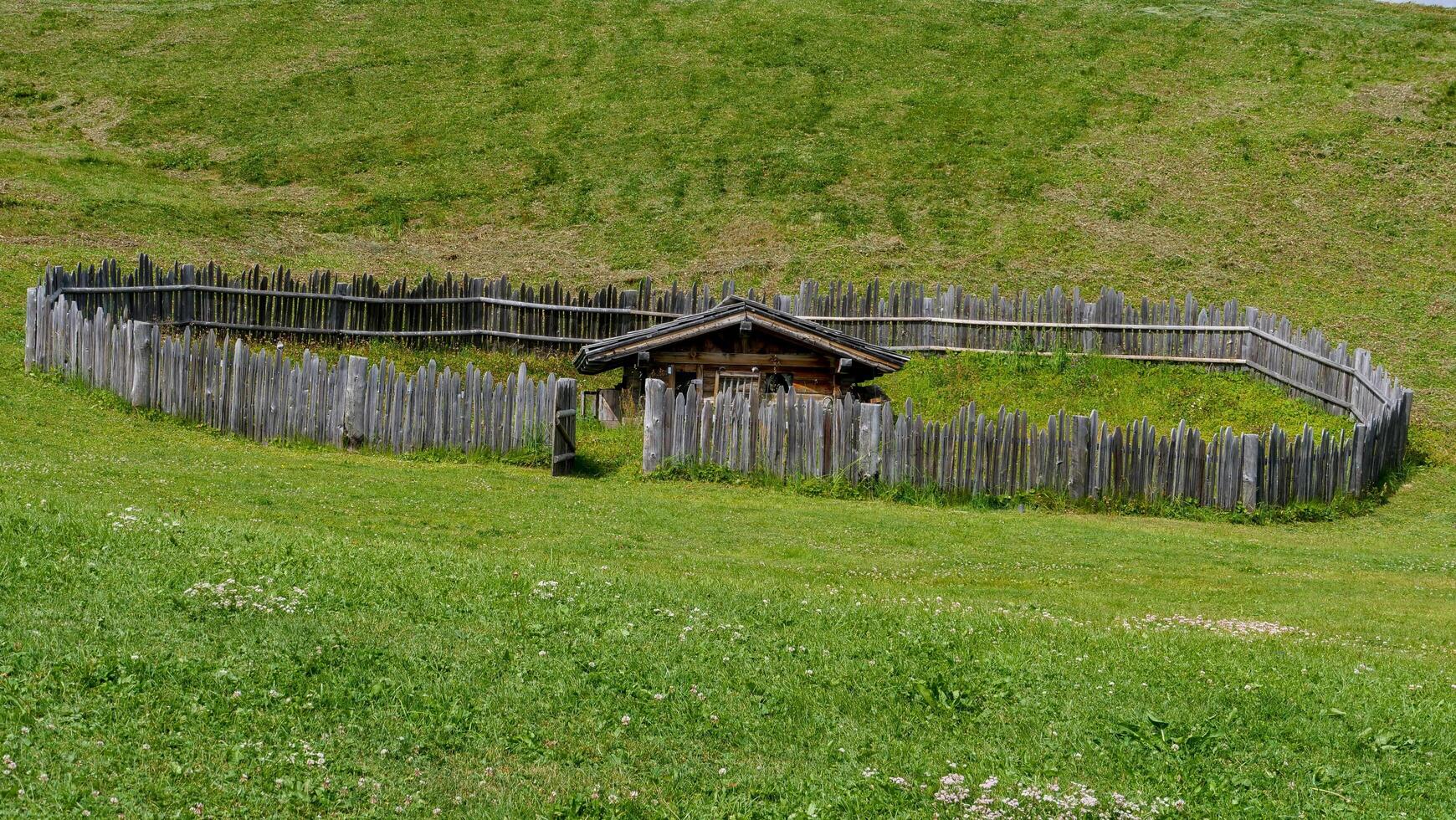 Historic wooden structure surrounded by a rustic fence on a sunlit grassy hillside in a tranquil rural setting photo