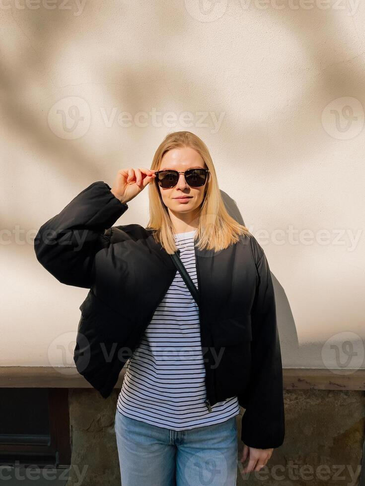 Stylish young blonde woman adjusting sunglasses while posing against white wall on sunny day, wearing black jacket, striped shirt, and jeans. Blonde woman in jeans, striped shirt, jacket, sunglasses photo