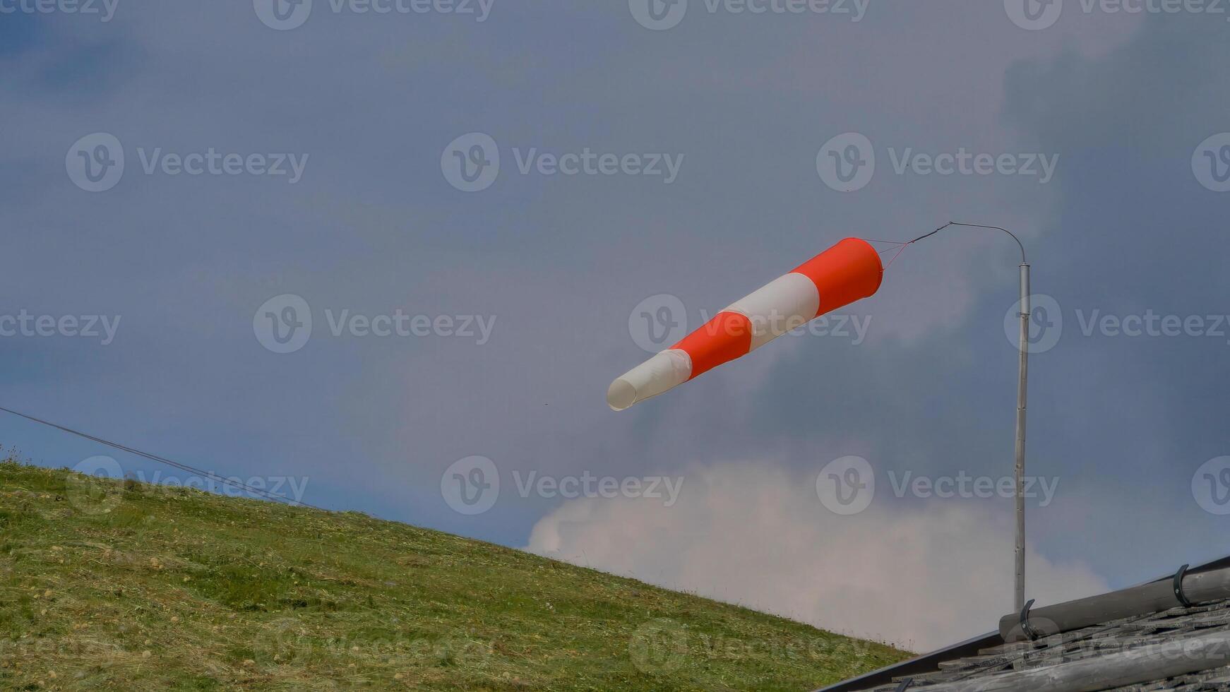 Wind direction indicator waves gracefully above a lush green hillside under a clouded sky in a serene rural setting photo