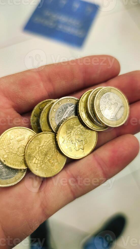 Hand holds various coins showcasing diverse currencies in a busy public space on a bright afternoon photo