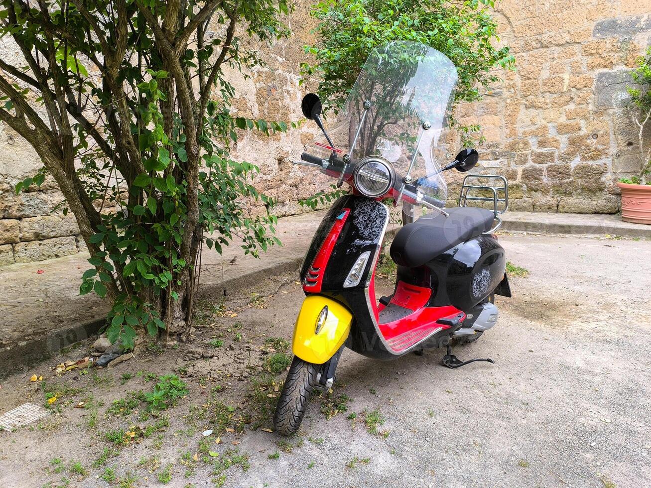 Brightly colored scooter parked under a lush green tree in a quaint cobblestone courtyard photo