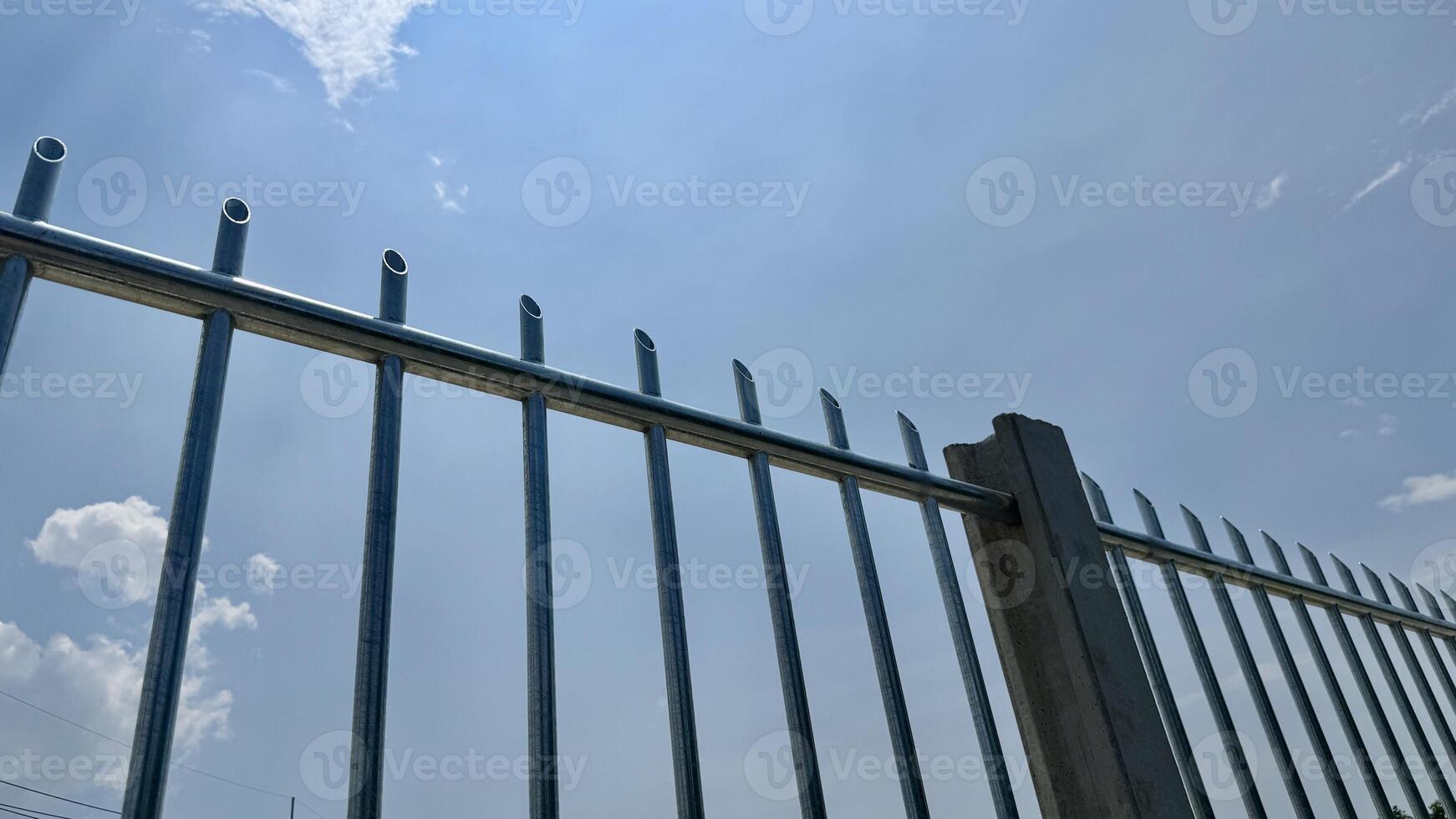Security Fence Under Blue Sky A close up view of a sturdy metal fence against a vibrant blue sky dotted with fluffy white clouds. The image evokes feelings of security protection and peace of mind. photo