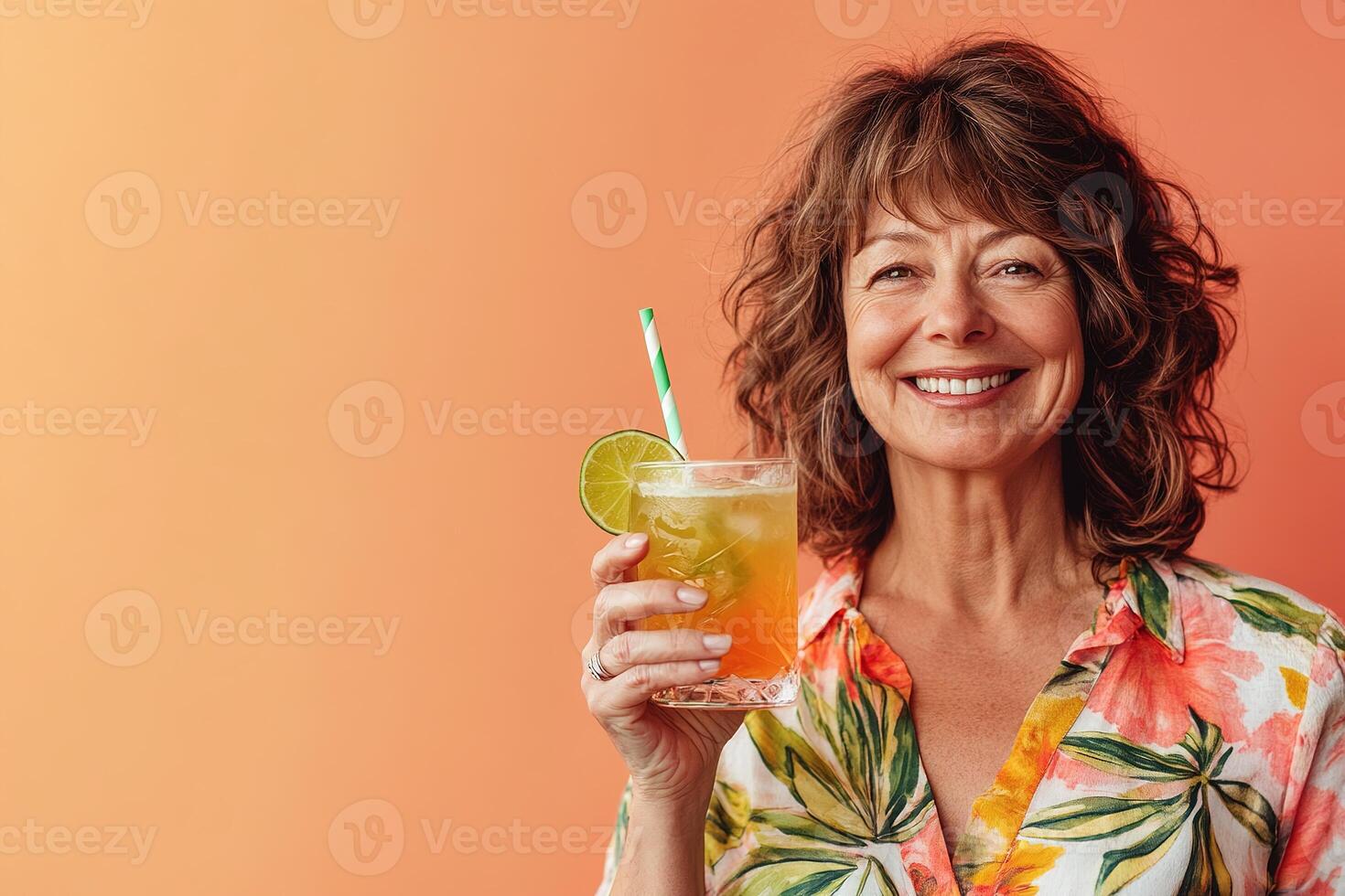 cheerful middle-aged woman holding a tropical cocktail with a slice of lime, standing against a gradient sunset-colored background photo