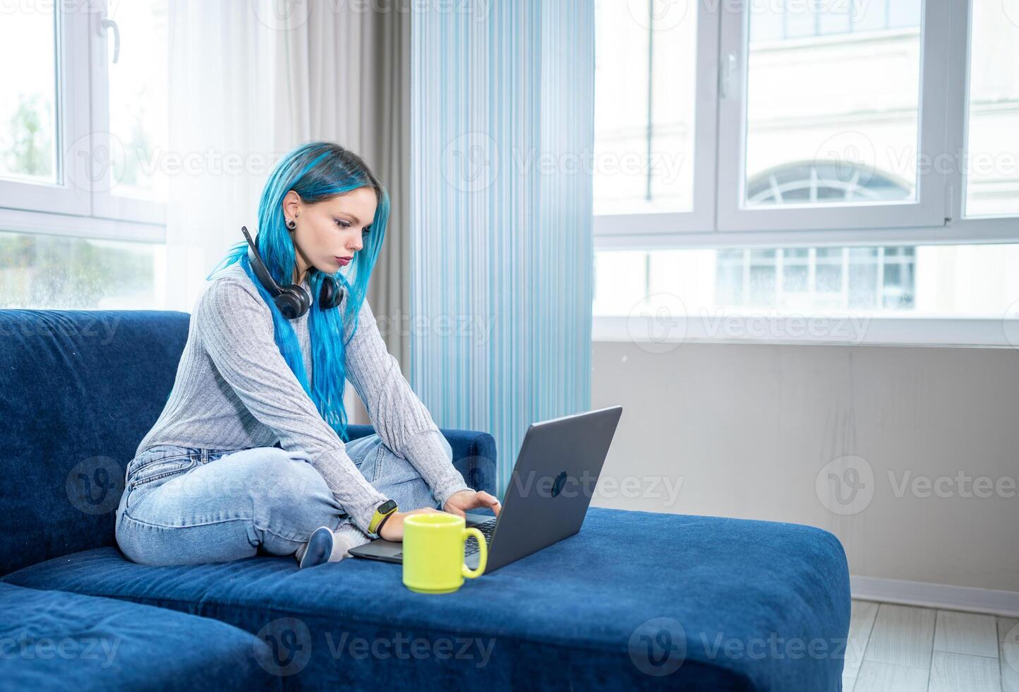 Beautiful blue haired woman working from home sitting on sofa, with headphones and microphone around her neck. Concept of working from home, using technology. photo