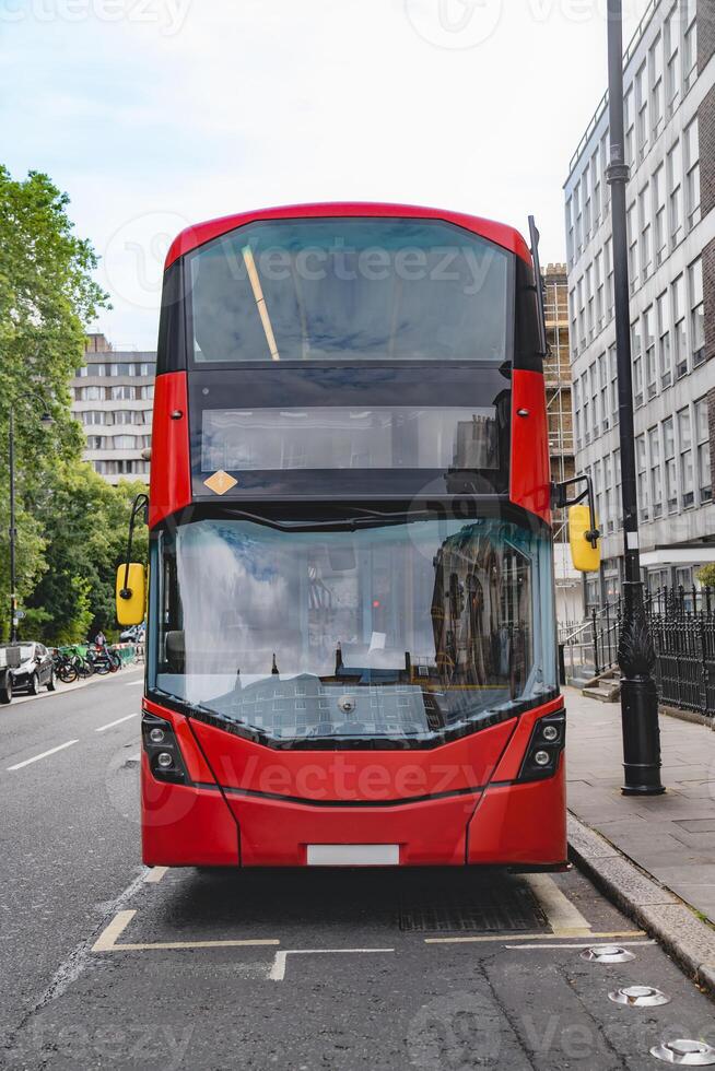 Front view of a red London double decker bus, showcasing the sharp design and clean lines. The surrounding urban landscape and buildings reflect in the bus windows. photo