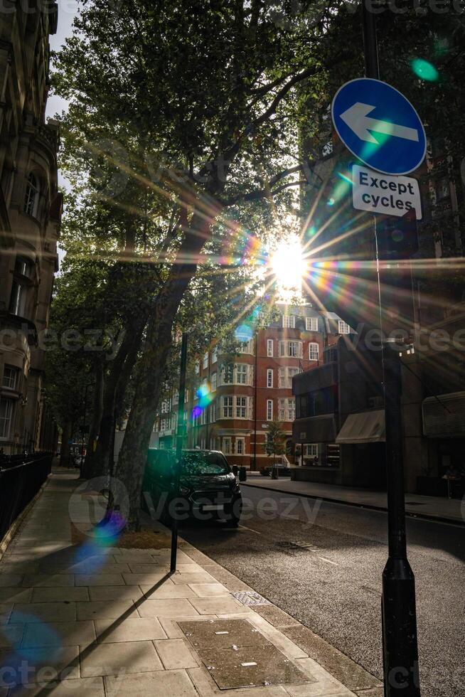 Sunlight shines through trees along a city street, with a road sign indicating a left turn for cycles. The urban scene features a quiet street with a parked vehicle a photo