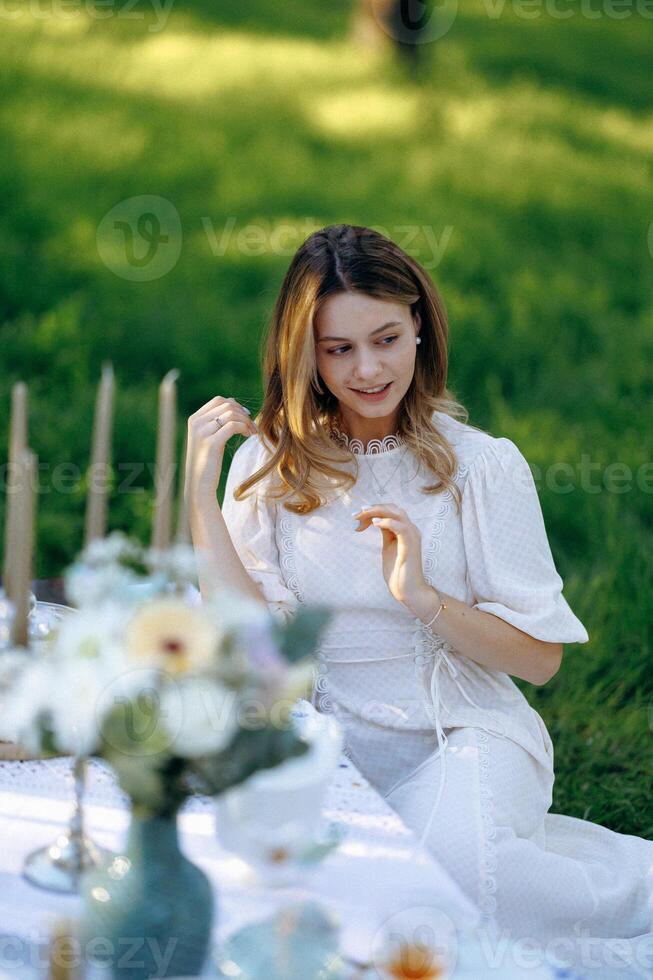 A woman in a white dress sits at a table in a serene setting photo