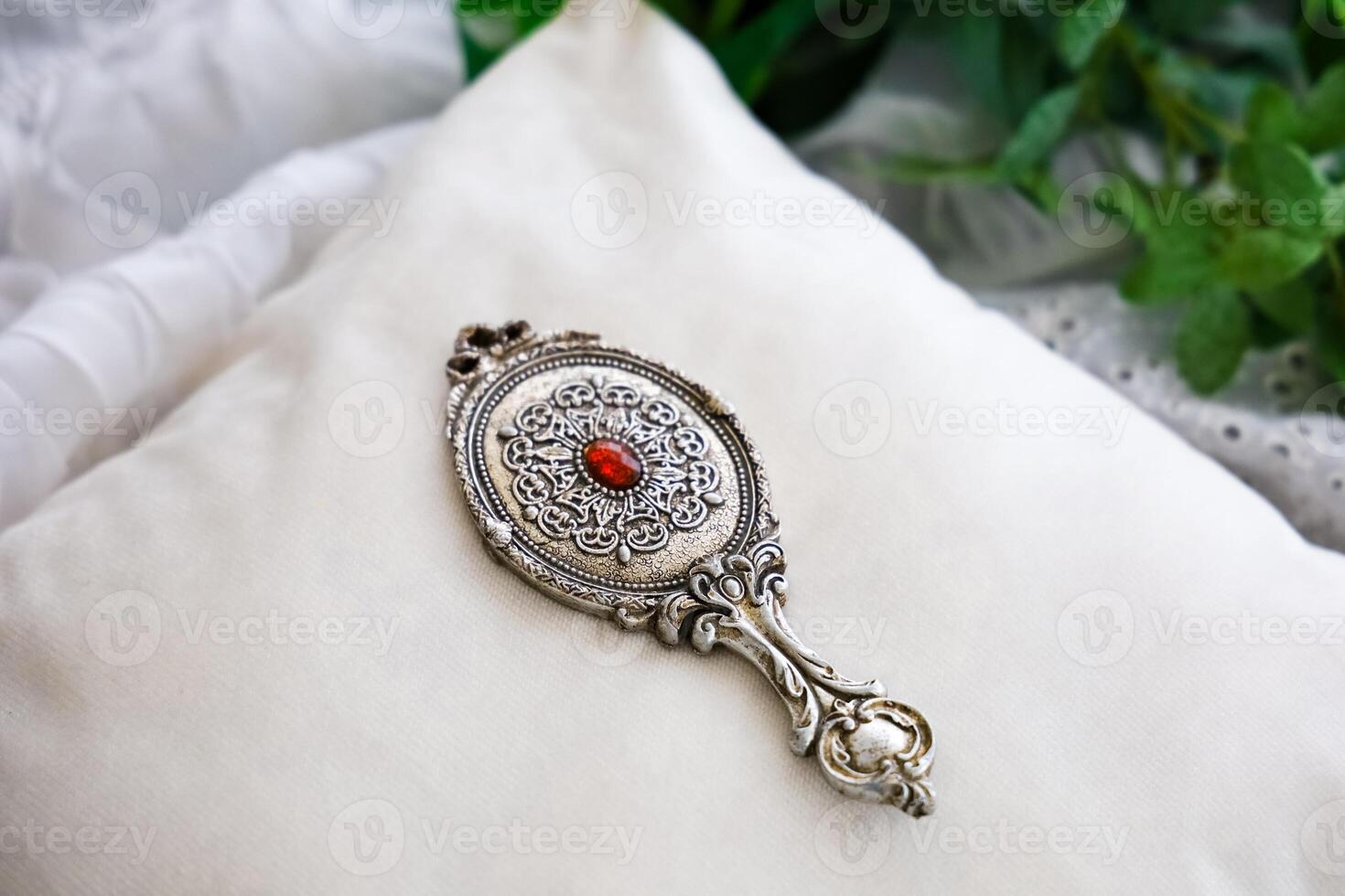 Detailed ornate silver hand mirror with red gem on a soft pillow surrounded by greenery photo