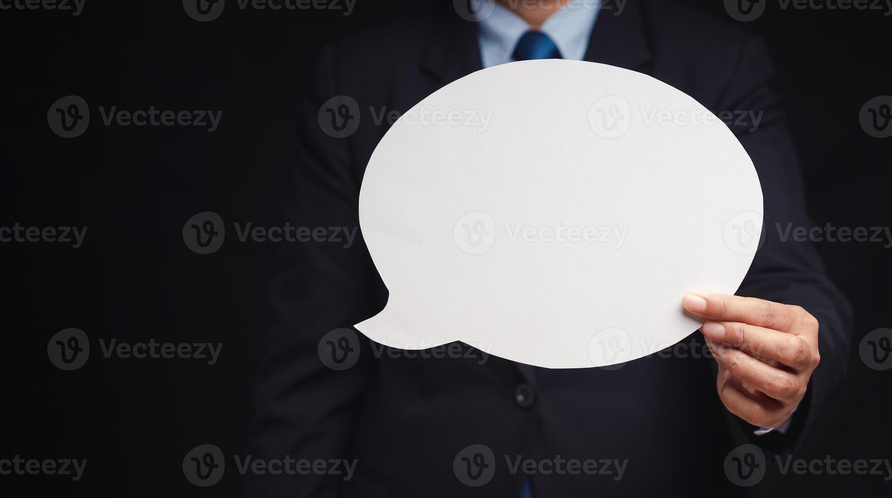 Close-up of a businessman in a suit holding a blank white speech bubble while standing on a black background. Space for text. photo