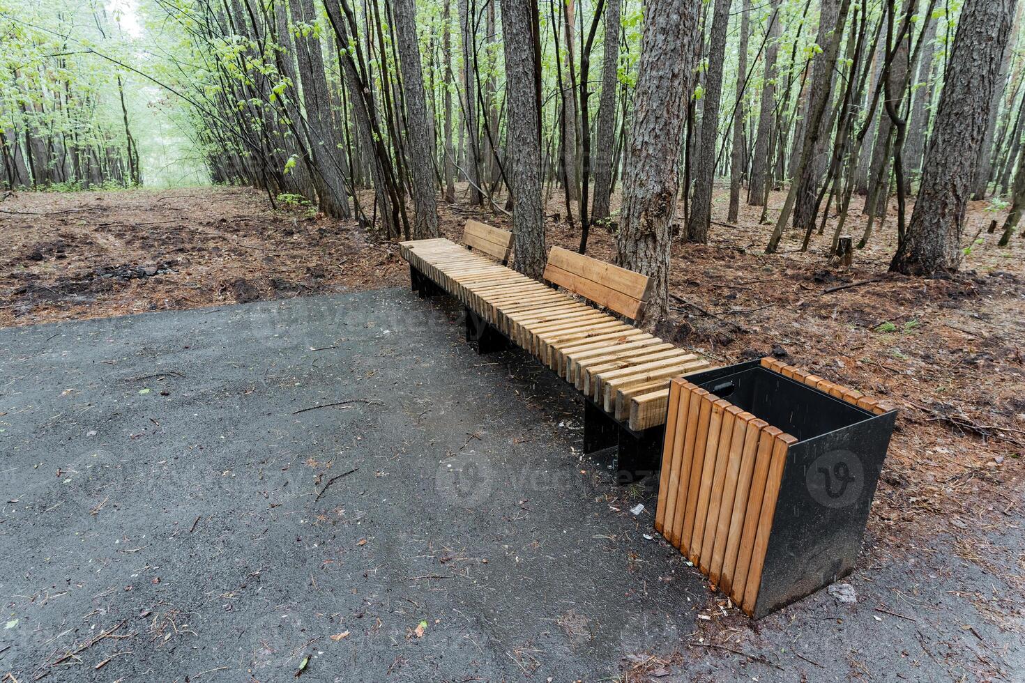 A strong wooden bench next to a trash can in a tranquil forest is the perfect spot to sit, relax, and admire the breathtaking beauty of nature, making it an ideal retreat photo
