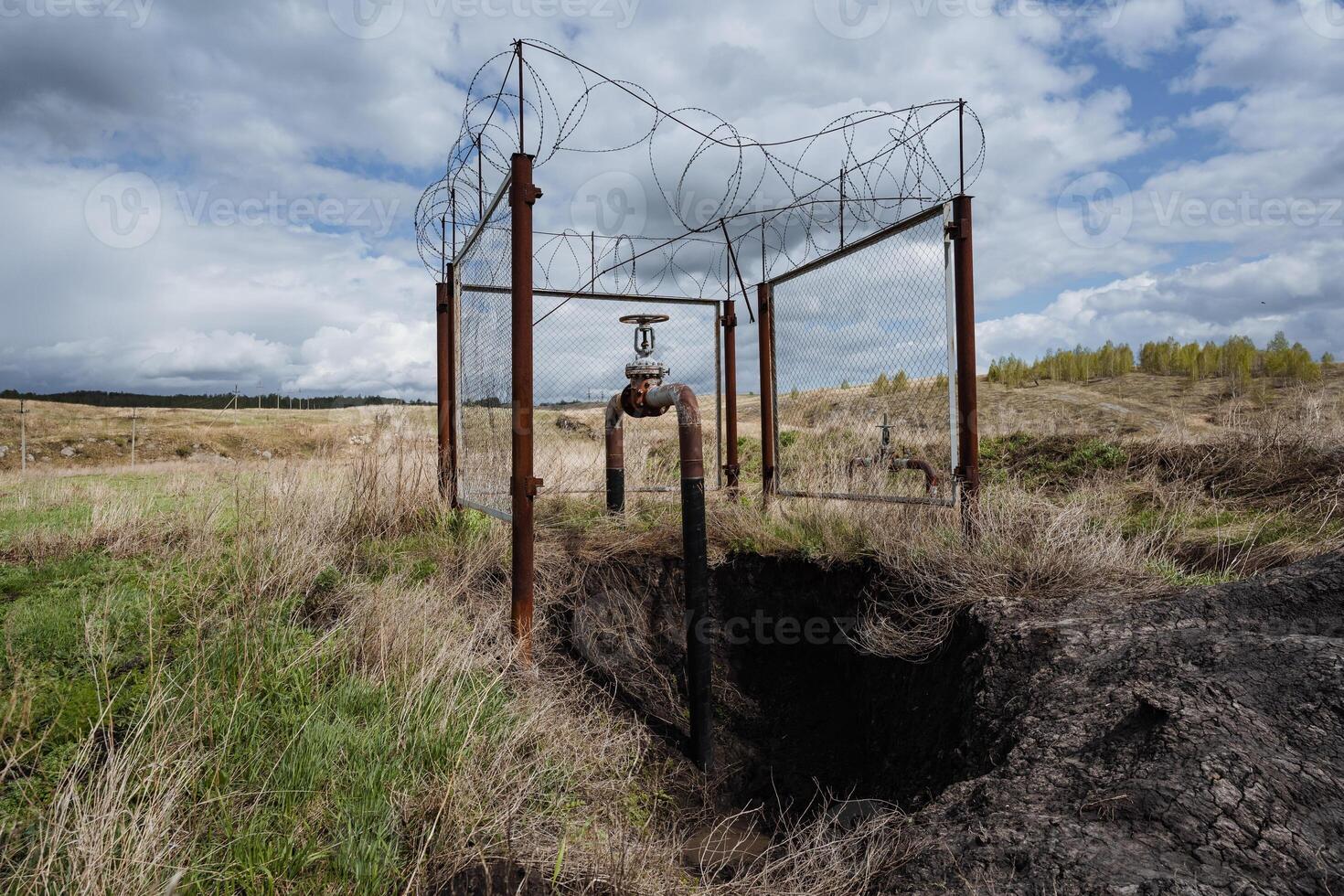 Abandoned Infrastructure in a Rural Landscape A disused rustic pipeline entwined with a barbed wire structure stands starkly against the backdrop of an empty field framed by the vast sky above photo