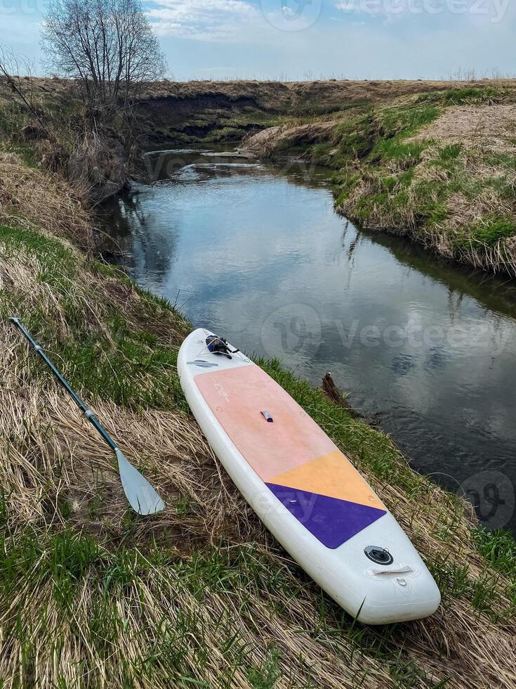 A paddle board along with a paddle is positioned next to a gently flowing river, surrounded by the natural beauty of the landscape, ready for an exciting adventure on the water photo