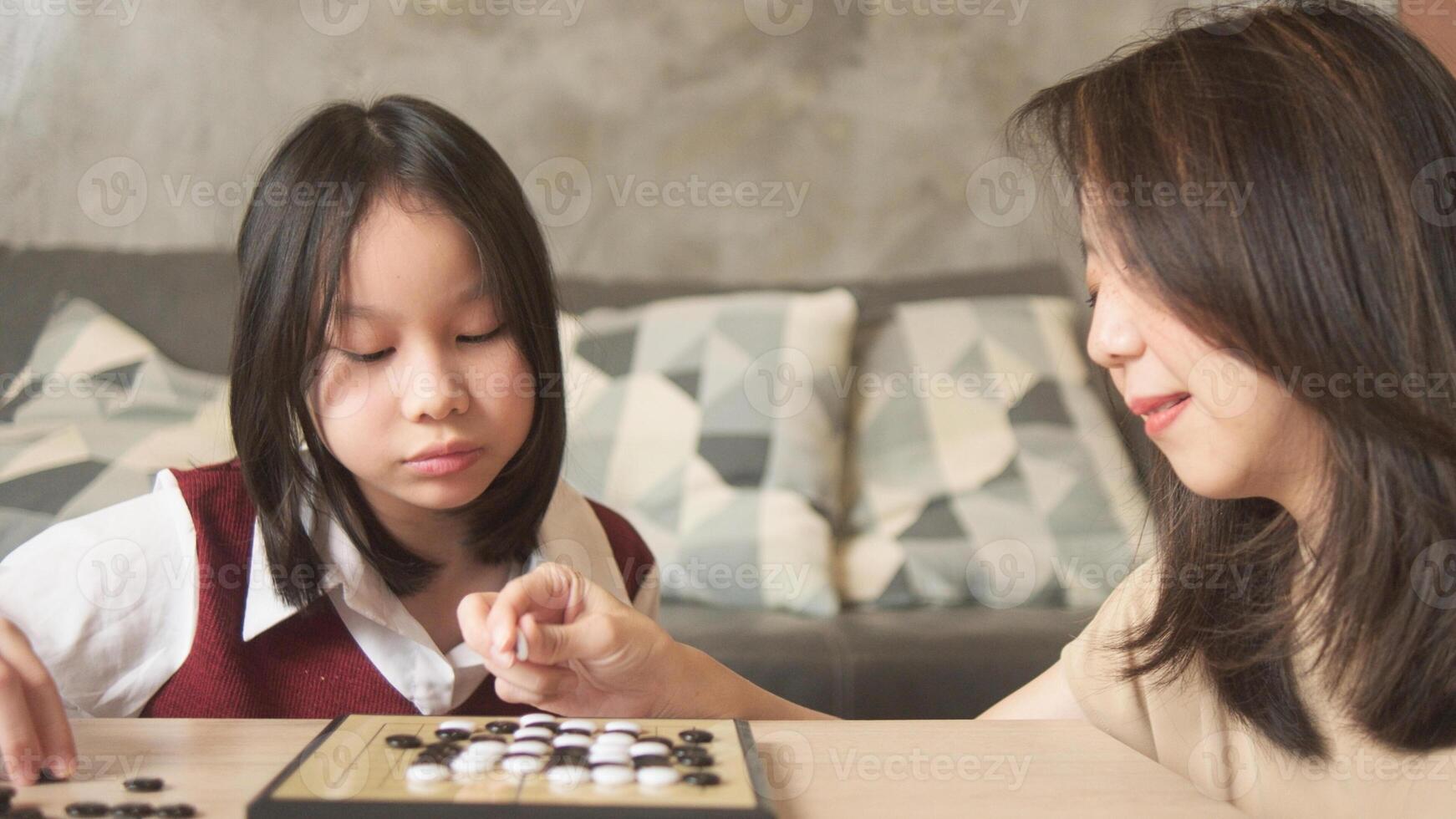 Asian family, child, and mother cheerful playing Go, checkers board game on table in home living room together, happy weekend relaxing hobby of kid and parent, concentrate and compete. photo