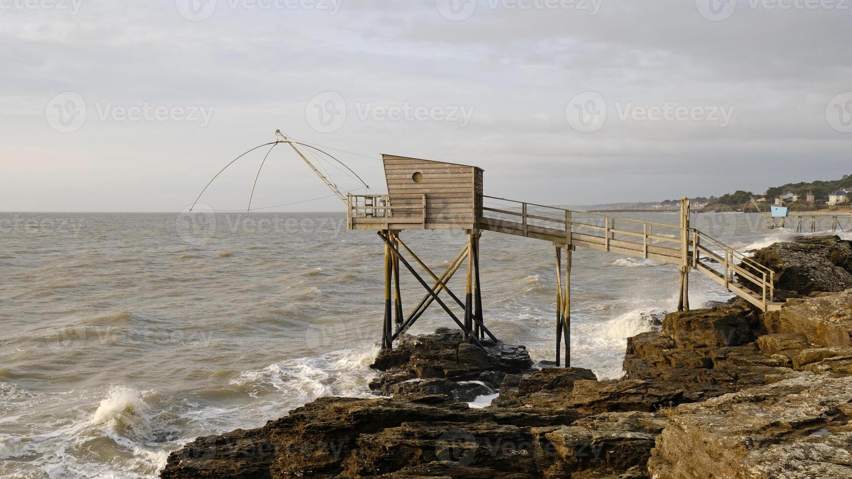 Traditional French fishing hut on stilts with carrelet net over ocean. Wooden structure stands on rocky shore, with waves and cloudy sky. Ideal for projects to travel photo
