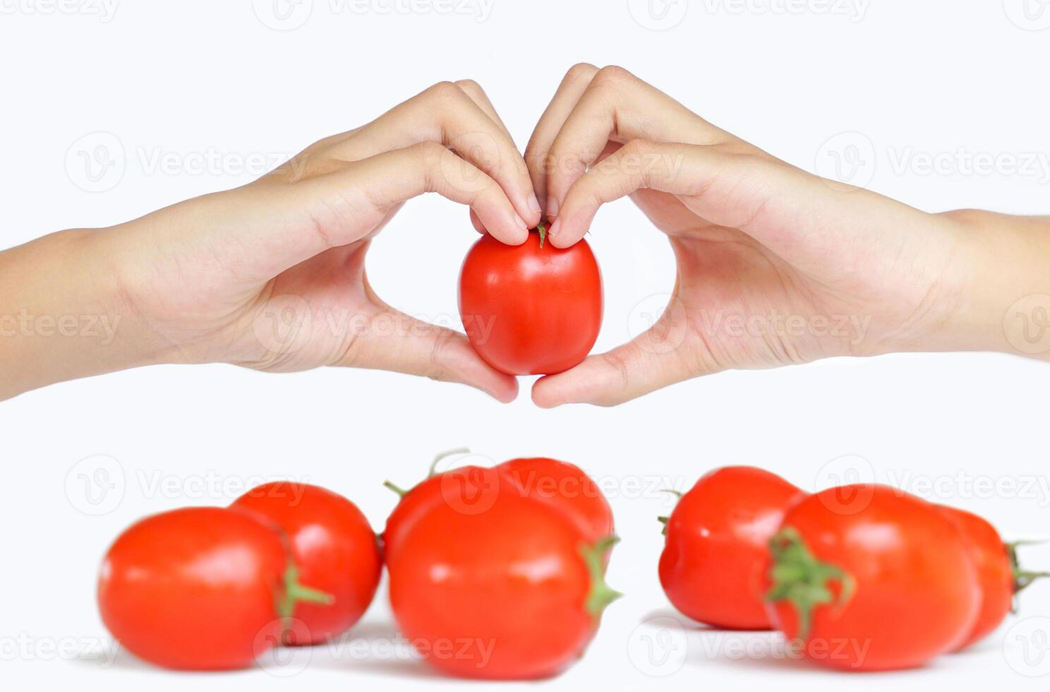 Focus on ripe tomato in hands clasped in heart shape on blur of bunch of tomatoes on white background, conveying the message of love for eating vegetables and tomatoes. photo