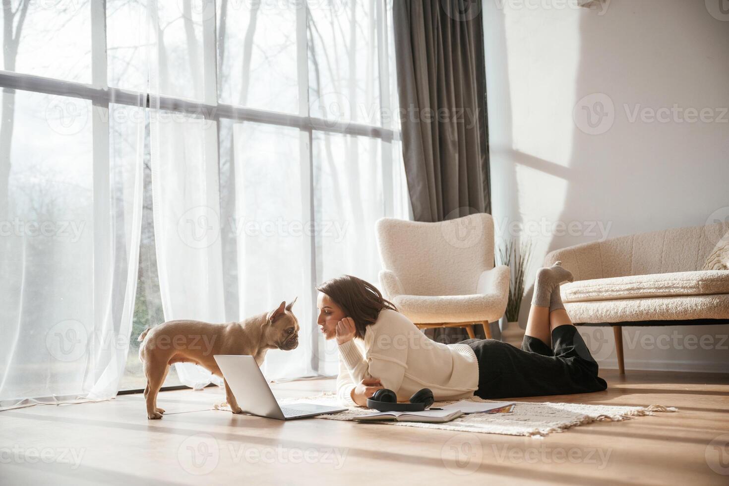 In, sunlight., Lying down on floor with laptop. Looking at pet. Young woman is with her pug dog at home photo