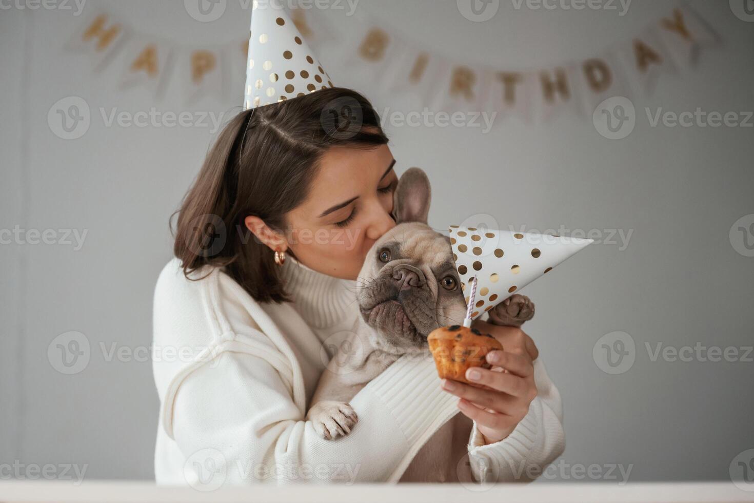 Happy owner is celebrating, birthday of pet. In hat and with cake. Young woman is with her pug dog at home photo