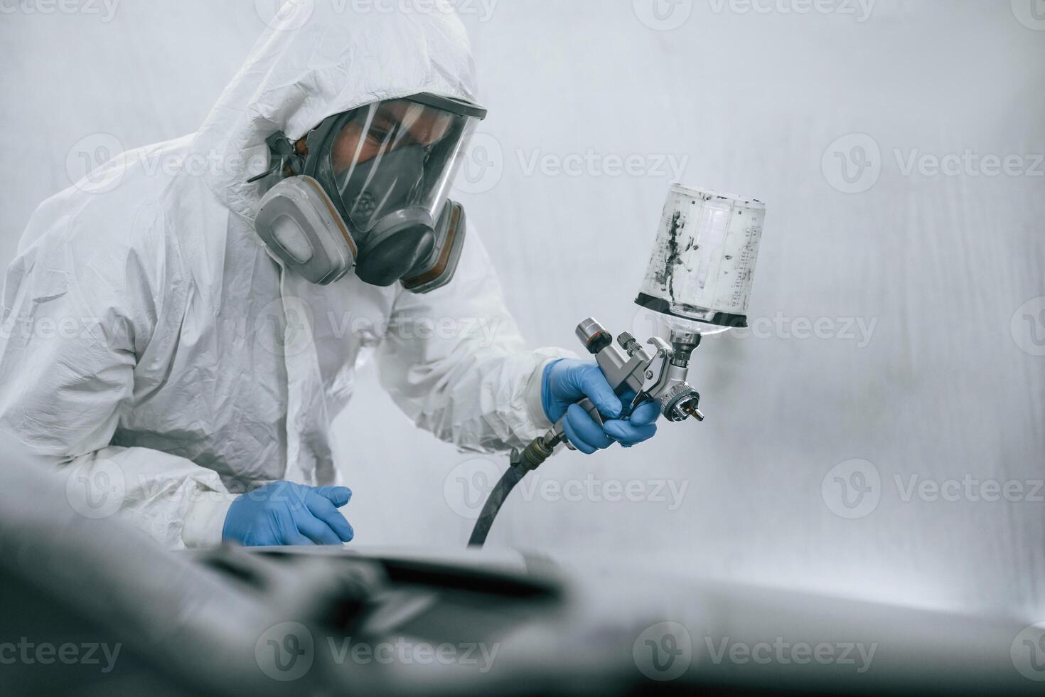 In protective mask,, and wear. Close up view of man that is painting a car by using spray guy photo