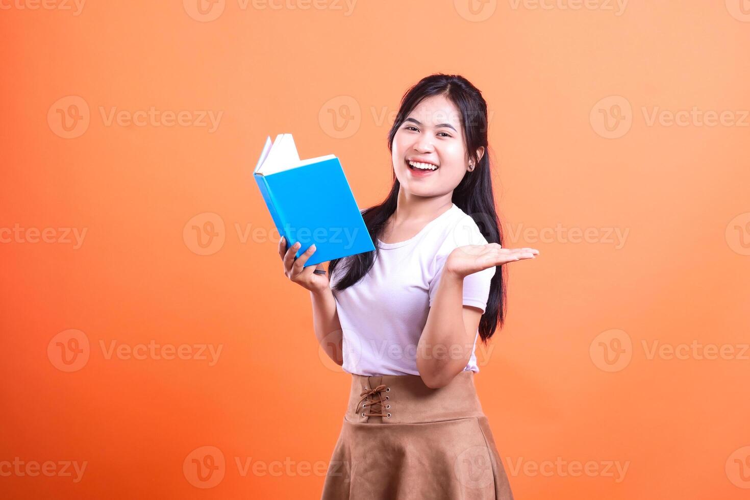 A woman holding a blue book and gesturing with her hand, standing against a vibrant orange background photo