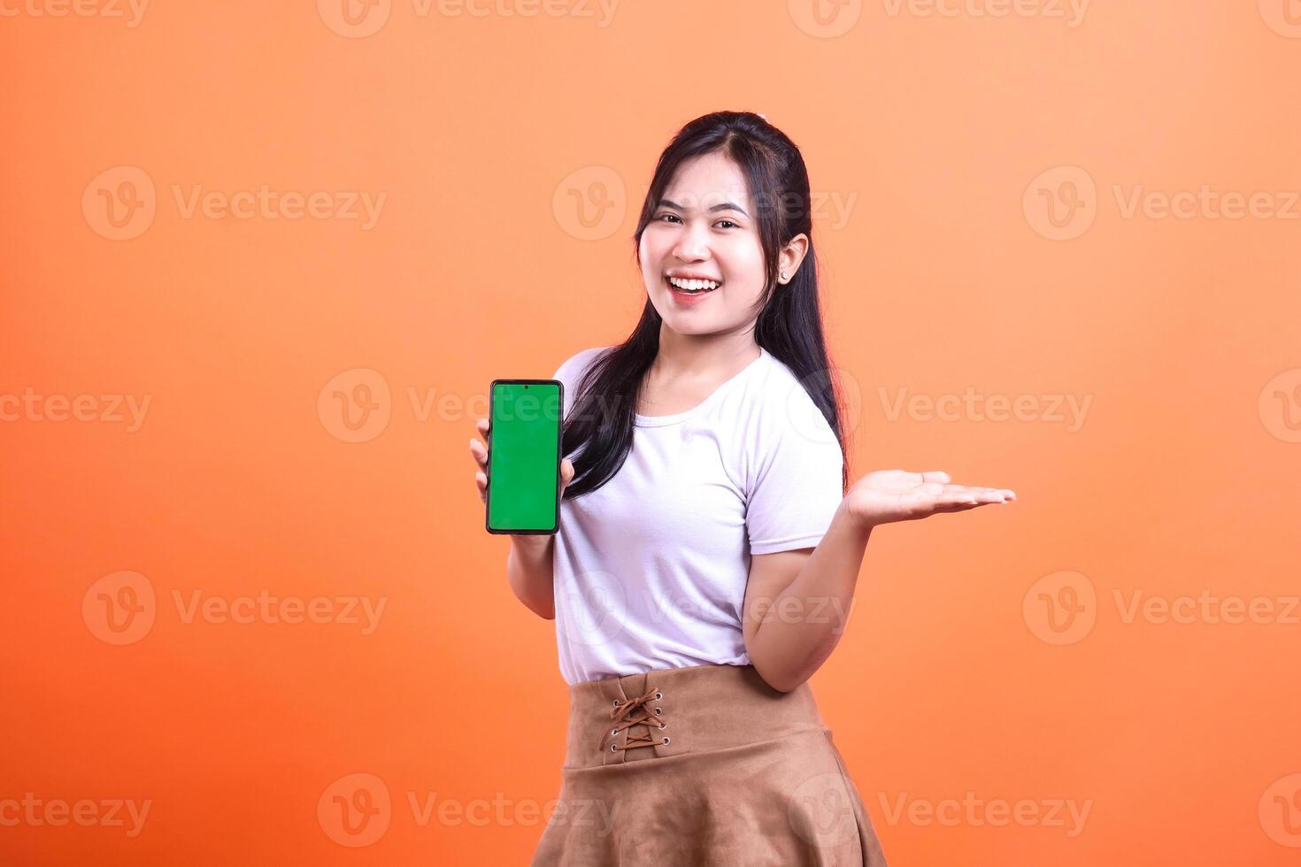Smiling woman holding green screen phone, showing something, isolated on orange background photo