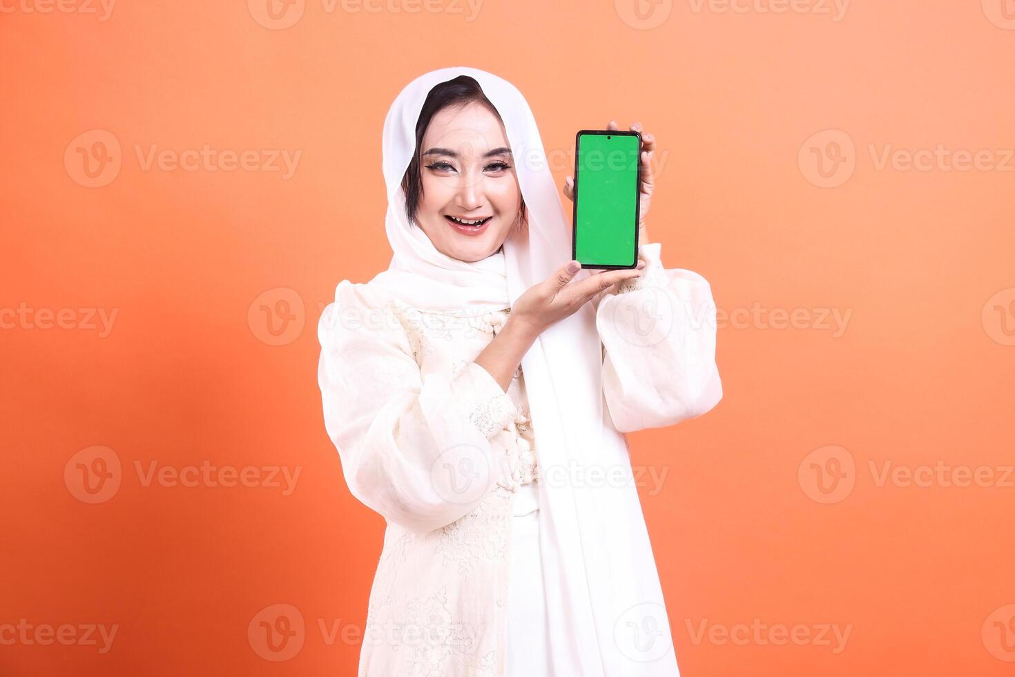 A smiling Muslim woman with a hijab is showcasing a smartphone with a green screen. Ideal for Ramadan-themed advertisements. isolated on a orange background. photo