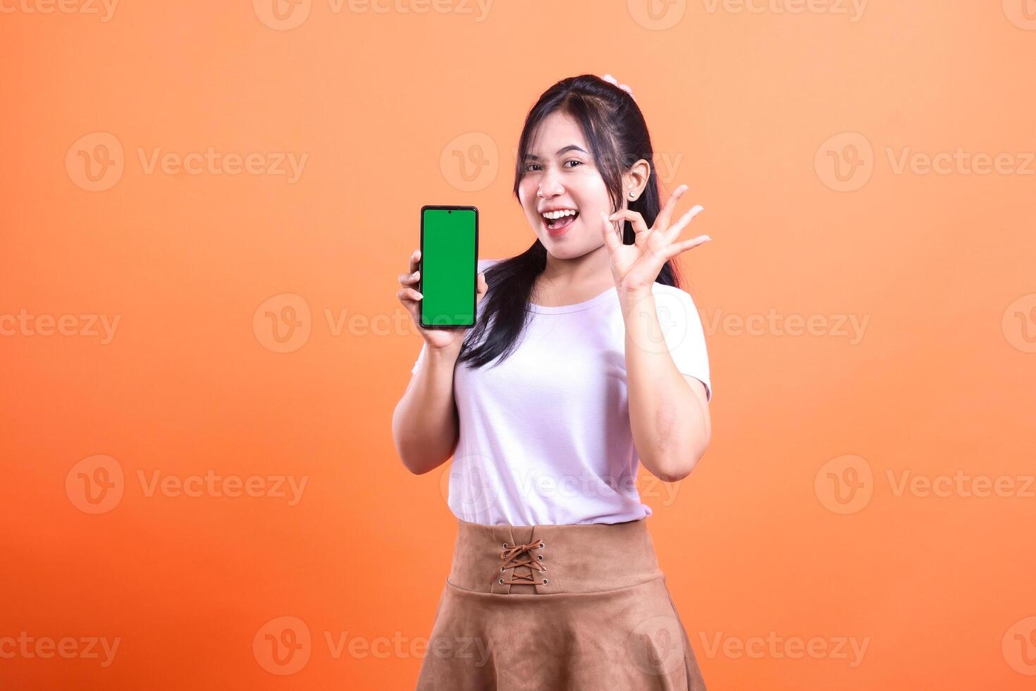 Smiling woman showing a smartphone with a green screen, okay hand gesture on orange background photo