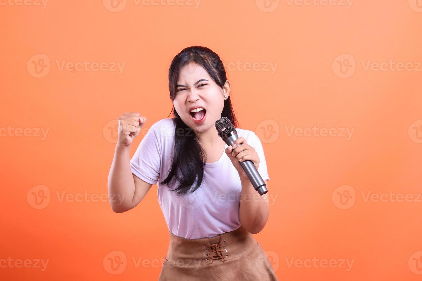 An energetic young woman sings passionately into a microphone, raising a fist in excitement while performing, isolated on orange background. photo