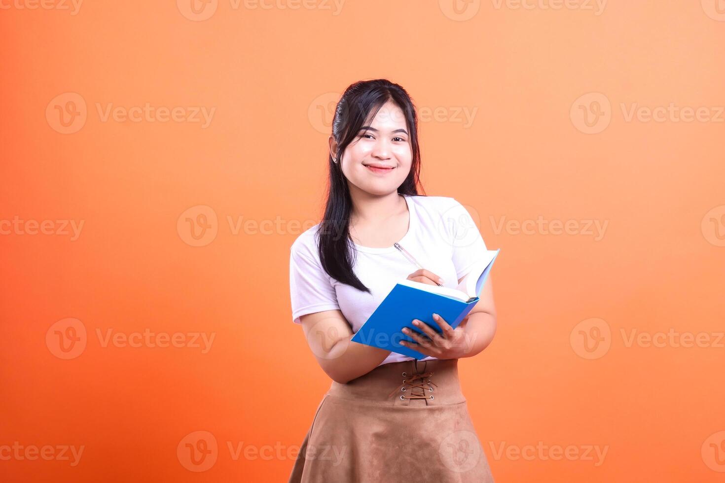A young woman is writing in a blue notebook with a pen while standing on a orange background photo