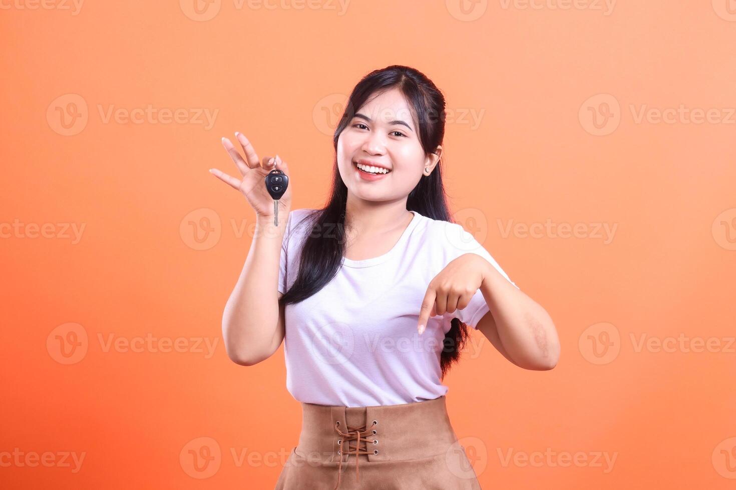 Smiling Asian woman holding car keys and pointing down at something exciting. This image is isolated on orange background. photo