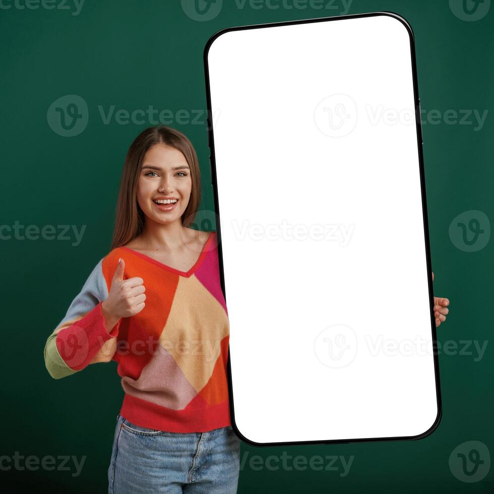 Large, smartphone. White display for text or design. Young woman is standing against green background in the studio photo