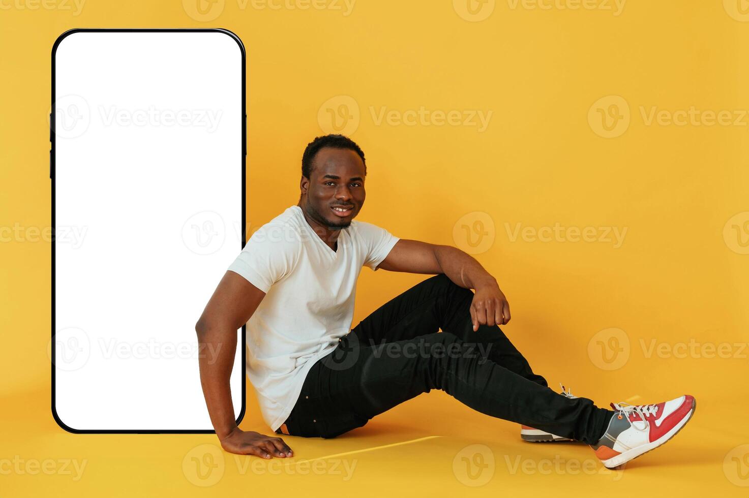 Sitting near to the ,,large ,smartphone with copy space. Black man is in the studio against yellow background photo