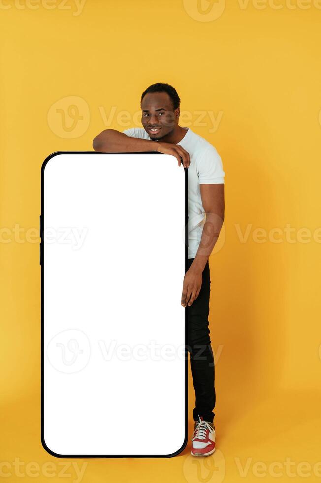 Leaning on the large smartphone ,for, copy space. Black man is in the studio against yellow background photo