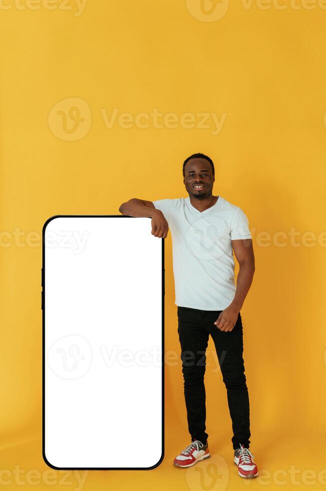 Leaning, on,, the large smartphone for copy space. Black man is in the studio against yellow background photo