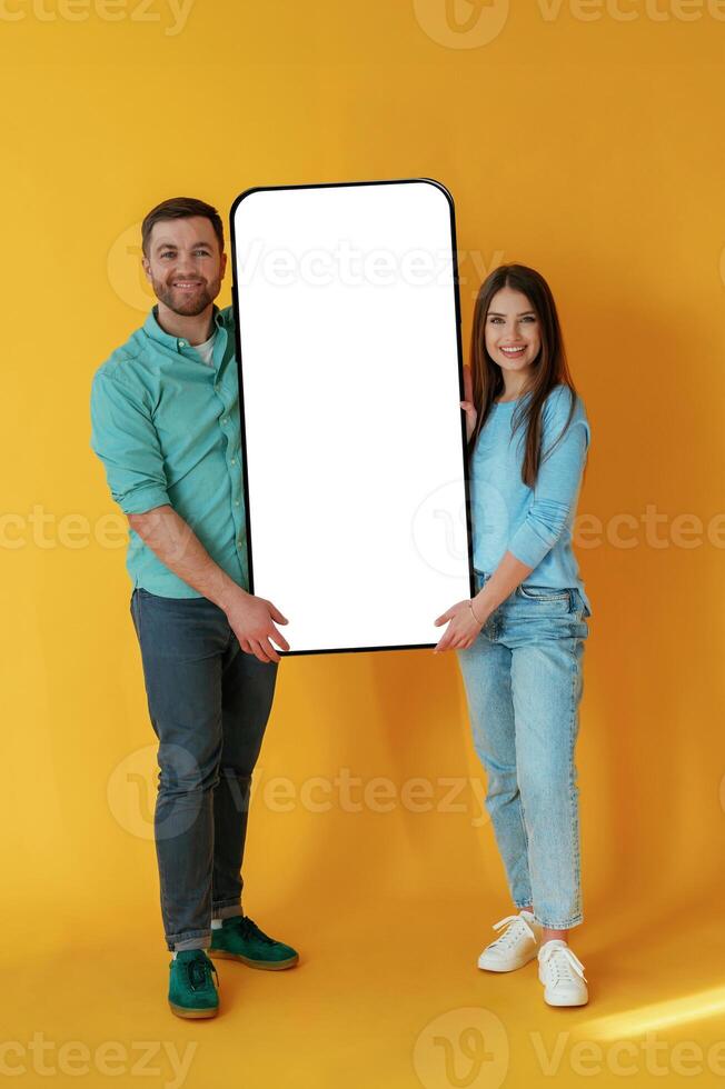 Holding ,white display, of smartphone, copy space. Man and woman are standing against yellow background photo