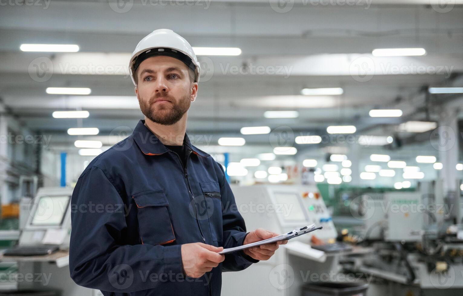 Looking far away, holding ,notepad. Factory, worker is indoors with hard hat photo
