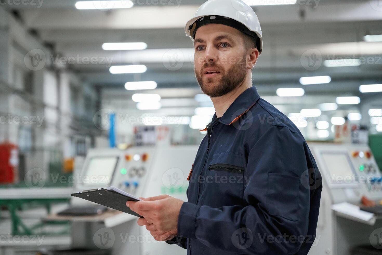 Looking far ,away, holding ,notepad. Factory worker is indoors with hard hat photo