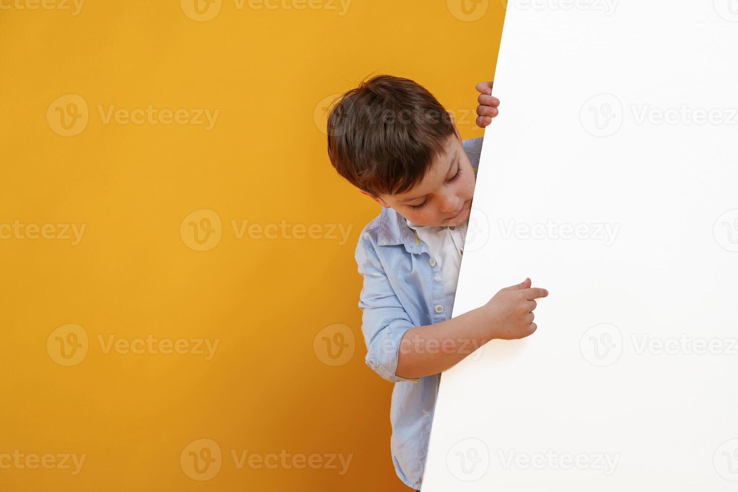 Showing something on the blank white surface. Little boy is in the studio against yellow background photo