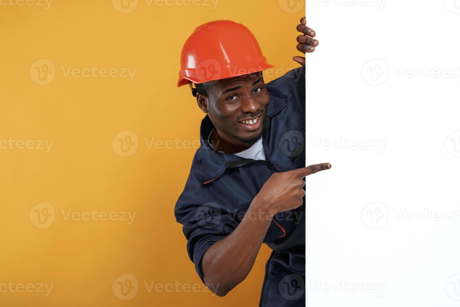 Engineer in ,hard hat is hiding behind white copy space. Handsome black man is in the studio against yellow ,,background photo