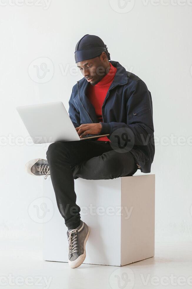 Sitting and using laptop. Black man in cap is in the studio against white background, photo
