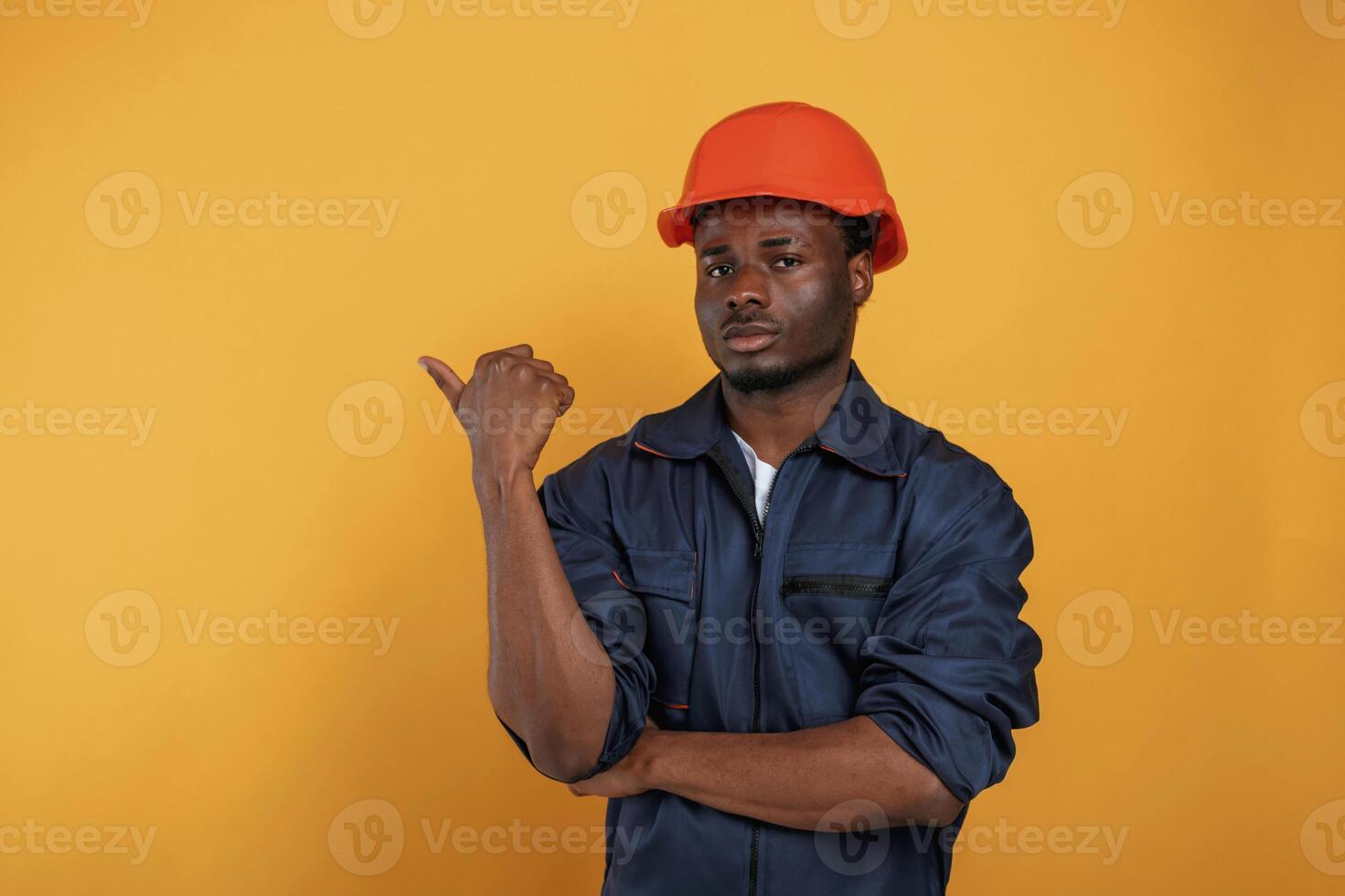 Guy in hard hat, showing ,copy,, space by finger. Handsome black man is in the studio against yellow background photo