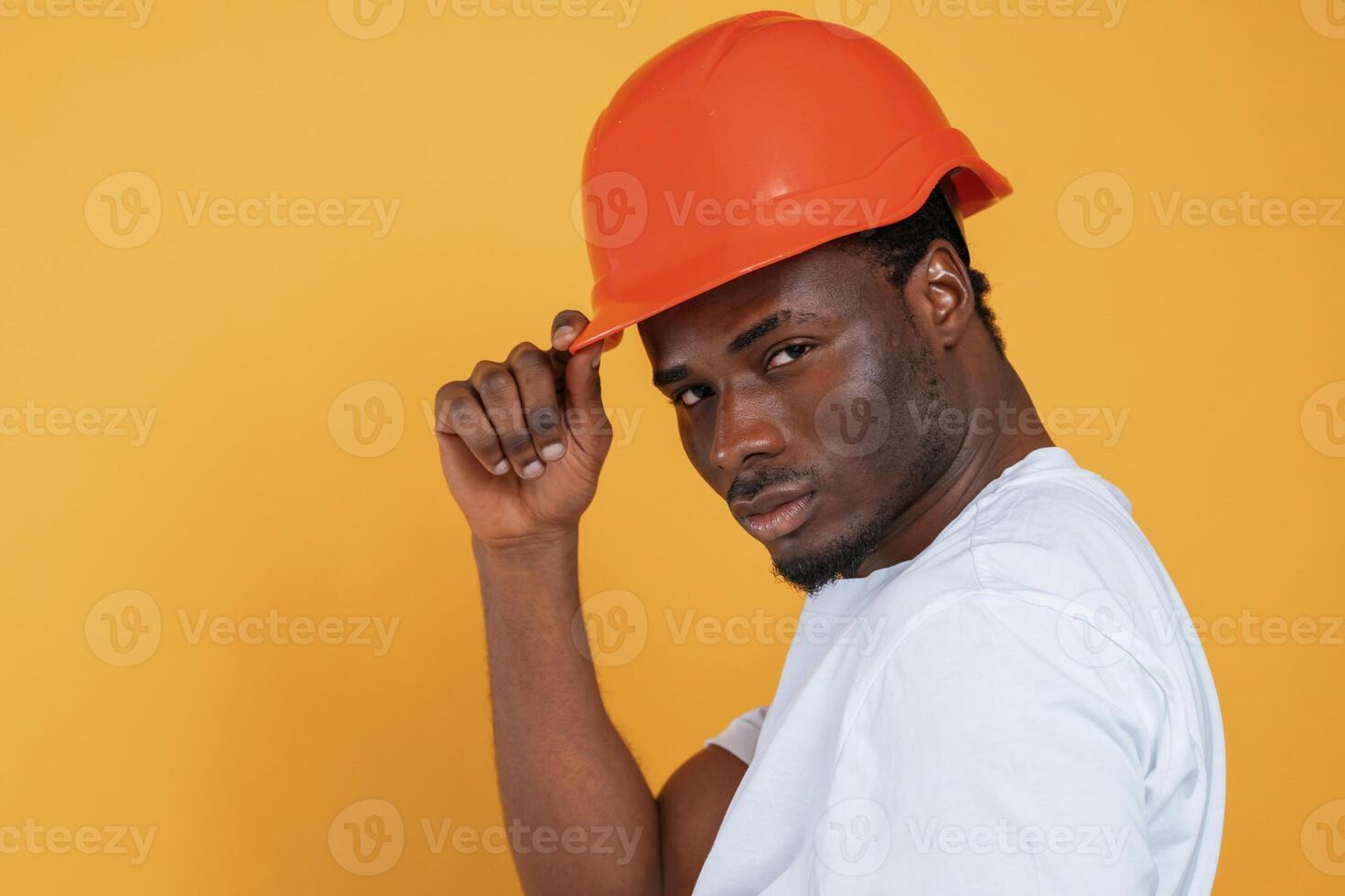 In orange hard hat. Construction worker. Handsome black man is in the studio against yellow background. photo