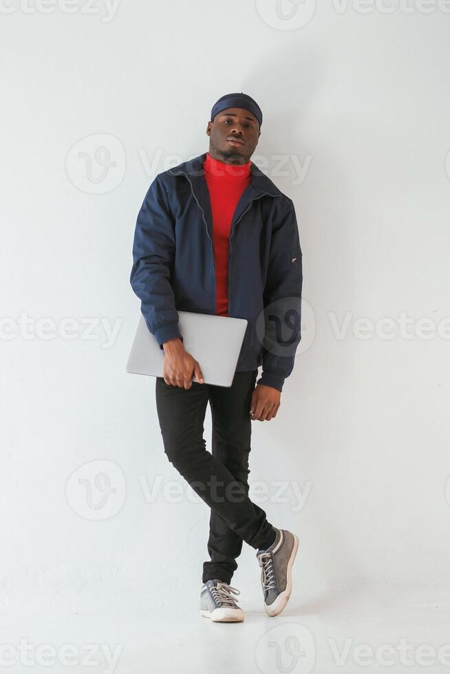 Standing and holding laptop in hand.., Black man in cap is in the studio against white background photo