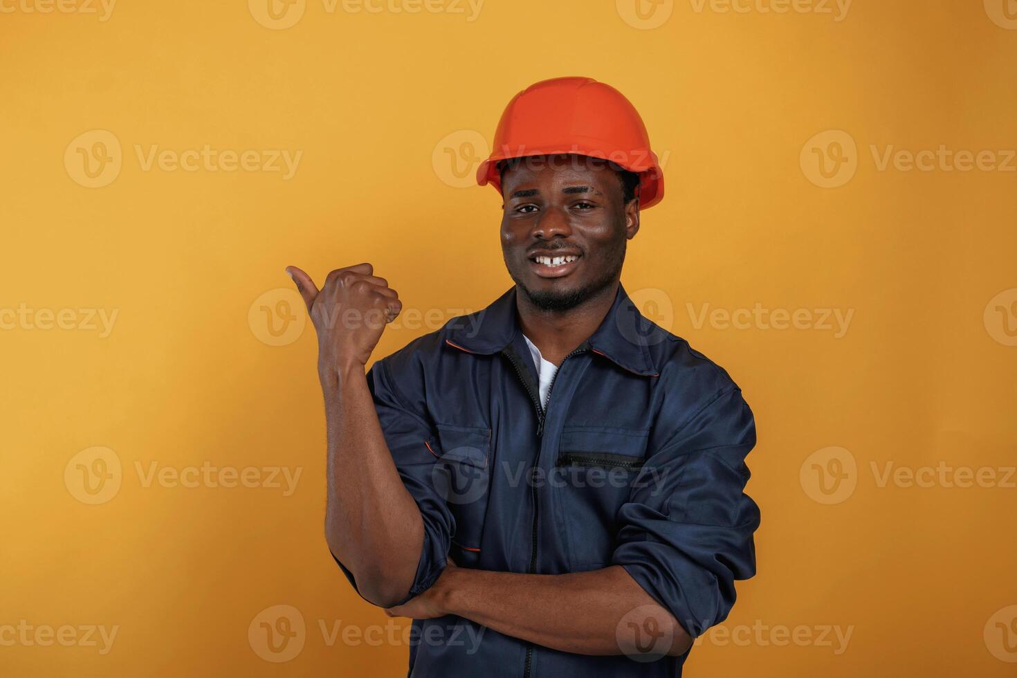 Guy, in ,hard, hat showing copy space by finger. Handsome black man is in the studio against yellow background photo
