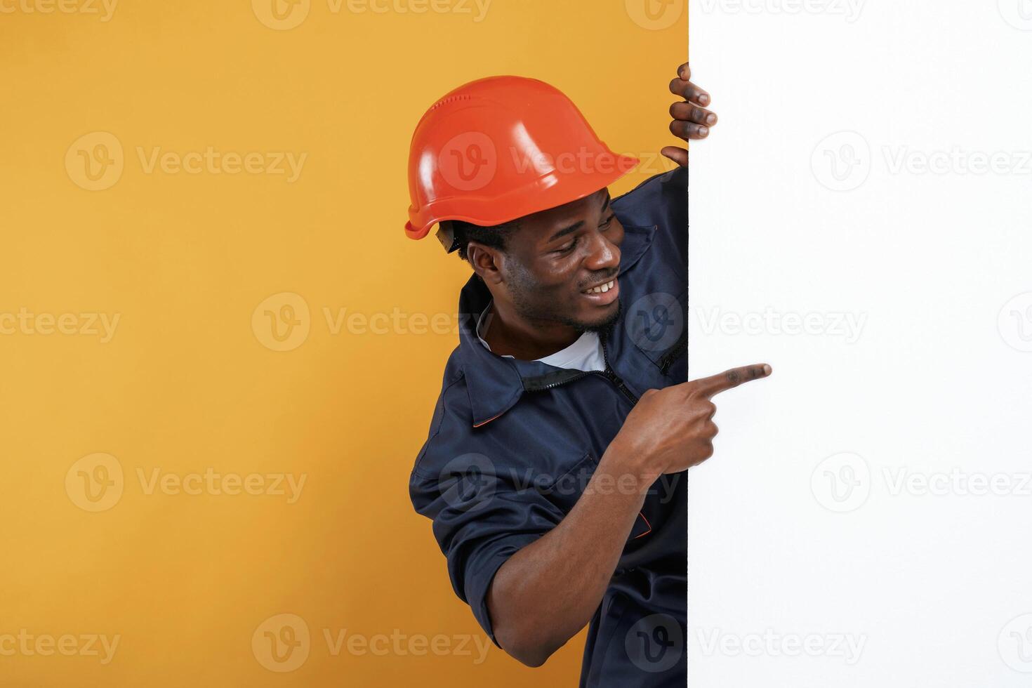 Engineer, in ,hard hat is hiding, behind white copy space. Handsome black man is in the studio against yellow background photo