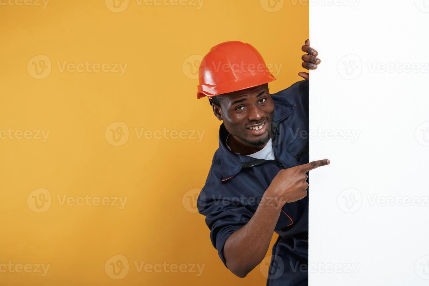 Engineer in hard hat is hiding behind, white copy space. Handsome black man is in the studio against yellow background photo