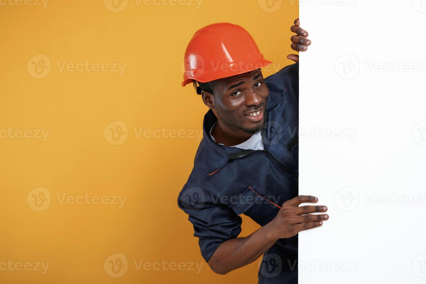 Engineer, in hard hat is hiding behind white copy space. Handsome black man is in the studio against yellow background photo