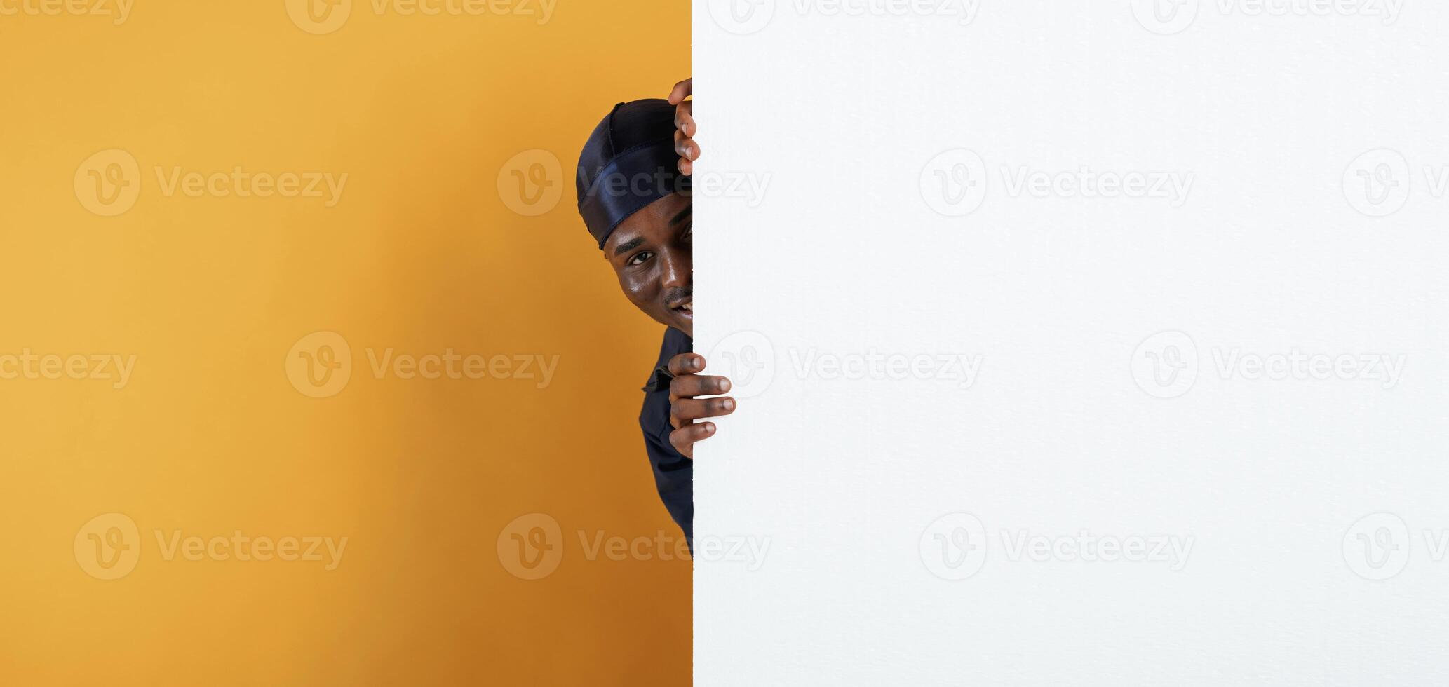 White, copy space, smiling and hiding behind it. Handsome black man is in the studio against yellow background photo